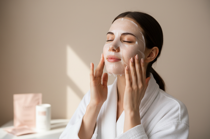 Photo portrait professionnelle d’une jeune femme avec une peau nette et lumineuse, en peignoir blanc, en train d’appliquer un masque visage en tissu collagène. Elle ajuste le masque avec ses mains sur ses joues, les yeux fermés, expression détendue. Fond uni beige clair, lumière naturelle douce, style campagne beauté skincare minimaliste, couleurs neutres et rose poudré, qualité photo de marque de cosmétique haut de gamme.
