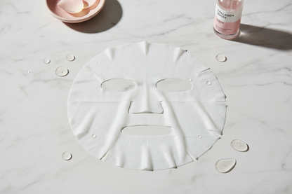 Photo professionnelle d’un masque visage en tissu collagène déplié, posé à plat sur une surface en marbre blanc avec quelques gouttes de sérum autour. Masque blanc légèrement brillant, texture bien visible, trous pour les yeux, le nez et la bouche. Ambiance très clean et skincare, lumière douce, style photo e-commerce haut de gamme, couleurs neutres avec des touches de rose poudré en décor.